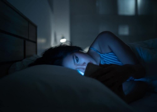 Женщина лежит в постели и смотрит в свой смартфон, а на ее лицо падает синий свет